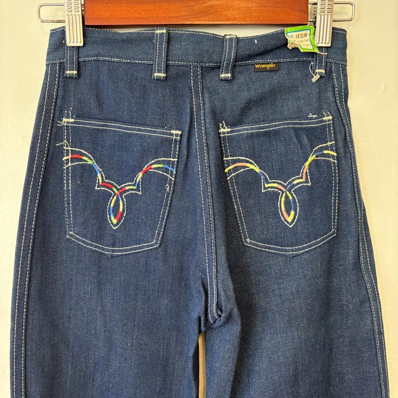 May include: Dark blue denim Wrangler jeans with white stitching and decorative back pockets. The pockets feature a colorful, embroidered design. The jeans are labeled "14 Slim".