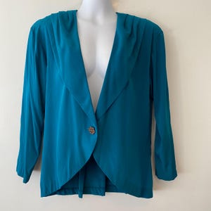 Könnte beinhalten: Türkisfarbener Blazer mit drapiertem Kragen und langen Ärmeln. Der Blazer hat einen einzelnen, dekorativen Knopfverschluss. Das Kleidungsstück wird auf einer Schaufensterpuppe präsentiert, die sein maßgeschneidertes Design und den fließenden Stoff zeigt.