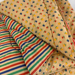 May include: A reversible quilted blanket featuring a tan background with a repeating pattern of small red, yellow, and blue tulip-like flowers on one side, and a pattern of horizontal stripes in red, blue, and green on the other side.