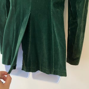 Puede incluir: Una chaqueta de terciopelo verde oscuro con mangas largas y un cierre de bot&oacute;n &uacute;nico. La chaqueta tiene una sola abertura en la espalda y una textura suave y lisa.