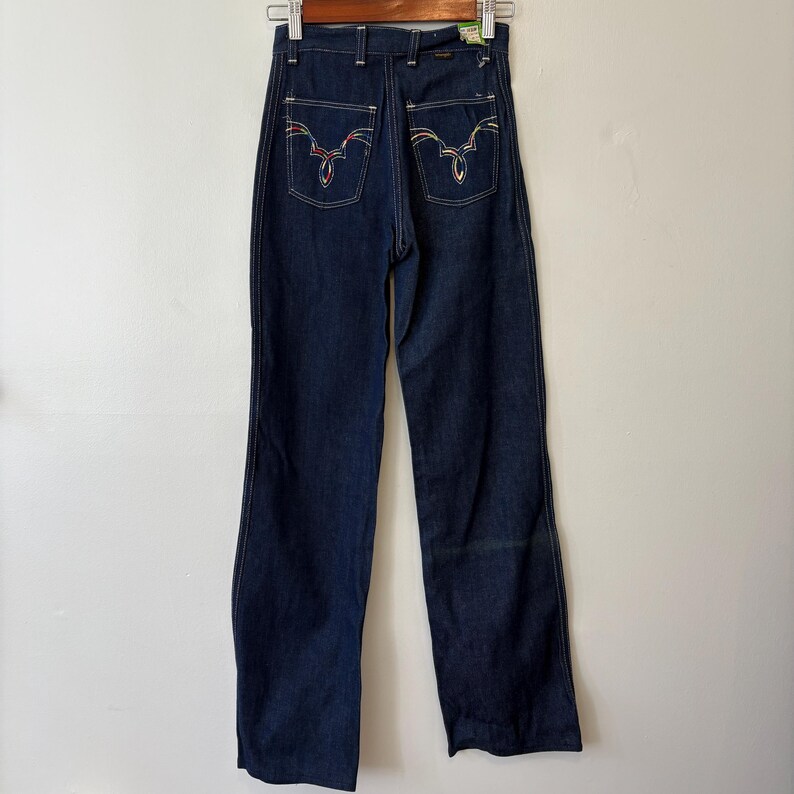 May include: Dark wash denim jeans with a high waist and wide leg. The back pockets feature colorful embroidered designs. The jeans are hanging on a wooden hanger.