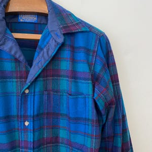 Pu&ograve; includere: Una camicia di flanella a quadri blu, verdi e viola con colletto e tasca blu. La camicia &egrave; abbottonata e ha un'etichetta in alto. Il tessuto ha un aspetto morbido e strutturato.