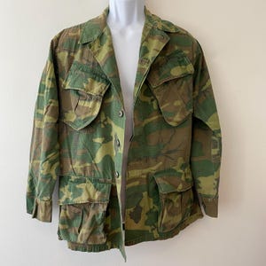 May include: A camouflage jacket in green, brown, and tan hues. It has multiple pockets, a button-up front, and a collared neckline. The jacket is displayed on a mannequin.
