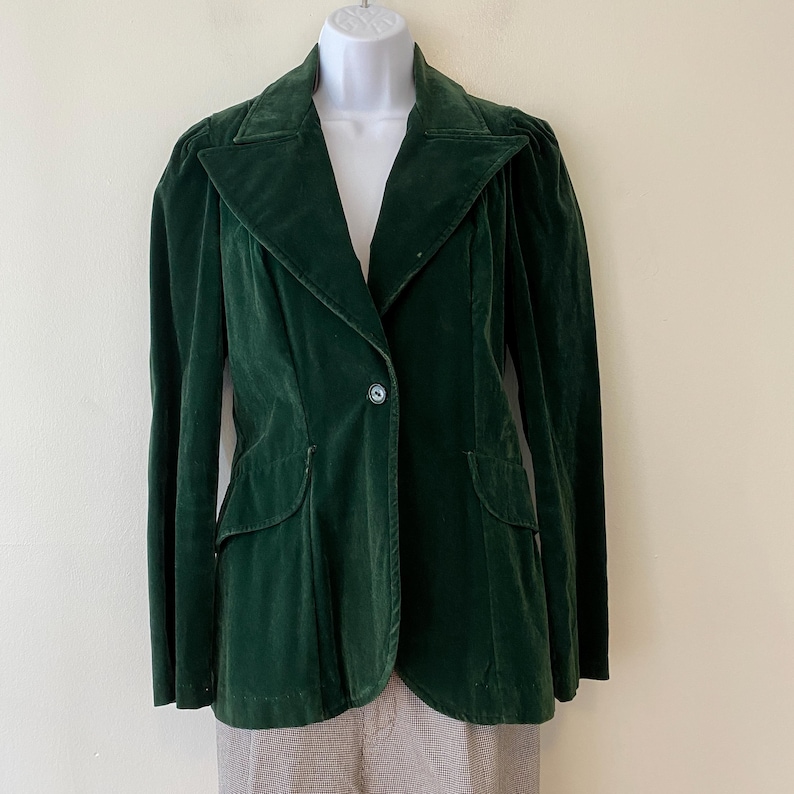 Puede incluir: Una chaqueta de terciopelo verde oscuro con un cierre de bot&oacute;n &uacute;nico y un cuello puntiagudo. La chaqueta tiene una silueta ajustada y presenta dos bolsillos en la parte delantera.