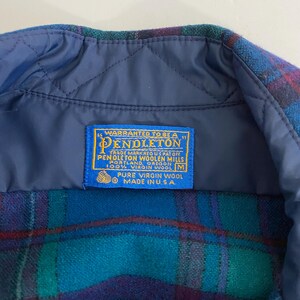 Pu&ograve; includere: Primo piano dell'etichetta di una camicia Pendleton a quadri blu, verdi e rossi. L'etichetta recita "WARRANTED TO BE A PENDLETON" e "PURE VIRGIN WOOL MADE IN U.S.A." La camicia &egrave; realizzata in 100% pura lana vergine.