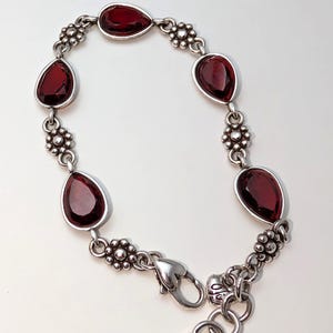 May include: Silver-toned bracelet featuring teardrop-shaped, deep red gemstones. The bracelet has a floral design between the gemstones and a heart-shaped clasp. A stylish piece of jewelry.