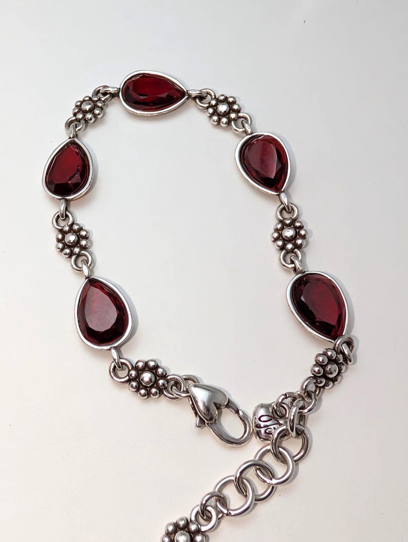 May include: Silver-toned bracelet featuring teardrop-shaped, deep red gemstones. The bracelet has a floral design between the gemstones and a heart-shaped clasp. A stylish piece of jewelry.