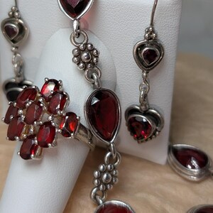 May include: A silver jewelry set featuring a ring, earrings, and a bracelet. The set includes oval and teardrop-shaped deep red gemstones. The earrings have heart-shaped accents. The ring features a cluster of oval gemstones.