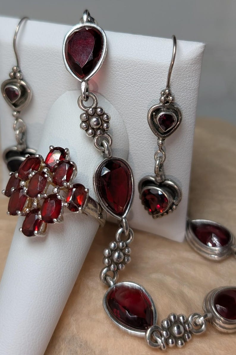 May include: A silver jewelry set featuring a ring, earrings, and a bracelet. The set includes oval and teardrop-shaped deep red gemstones. The earrings have heart-shaped accents. The ring features a cluster of oval gemstones.
