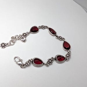 May include: Silver bracelet featuring teardrop-shaped, deep red gemstones. The bracelet has a floral design between each gem and a heart-shaped charm near the clasp. The bracelet is on a white background.