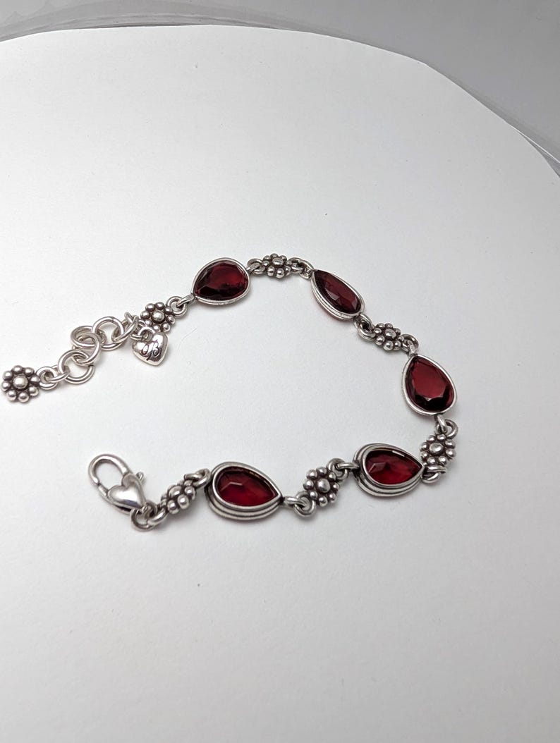May include: Silver bracelet featuring teardrop-shaped, deep red gemstones. The bracelet has a floral design between each gem and a heart-shaped charm near the clasp. The bracelet is on a white background.