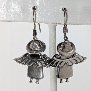 Op de afbeelding: Een paar zilverkleurige engelenoorbellen. Elke oorbel heeft een gestileerd engelontwerp met een halo, vleugels en een eenvoudige jurk. De oorbellen hangen aan een draadhaak met een spiraalvormig detail.