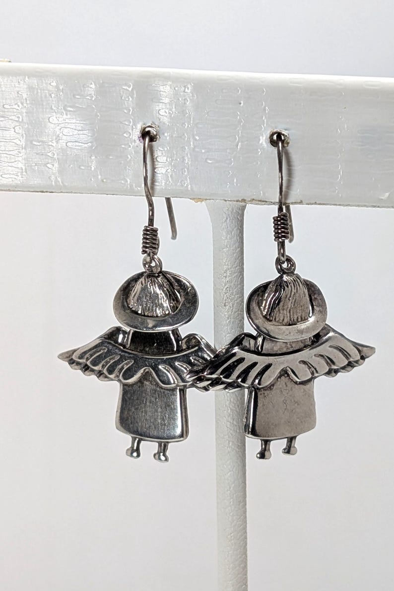 Op de afbeelding: Een paar zilverkleurige engelenoorbellen. Elke oorbel heeft een gestileerd engelontwerp met een halo, vleugels en een eenvoudige jurk. De oorbellen hangen aan een draadhaak met een spiraalvormig detail.