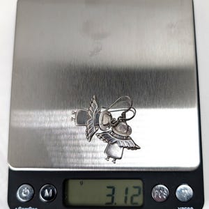 Op de afbeelding: Een paar zilverkleurige engelen oorbellen met gedetailleerde vleugels en hoeden, liggend op een digitale weegschaal. De weegschaal geeft een gewicht van 3,12 gram weer. De oorbellen zijn het middelpunt en tonen een grillig ontwerp.