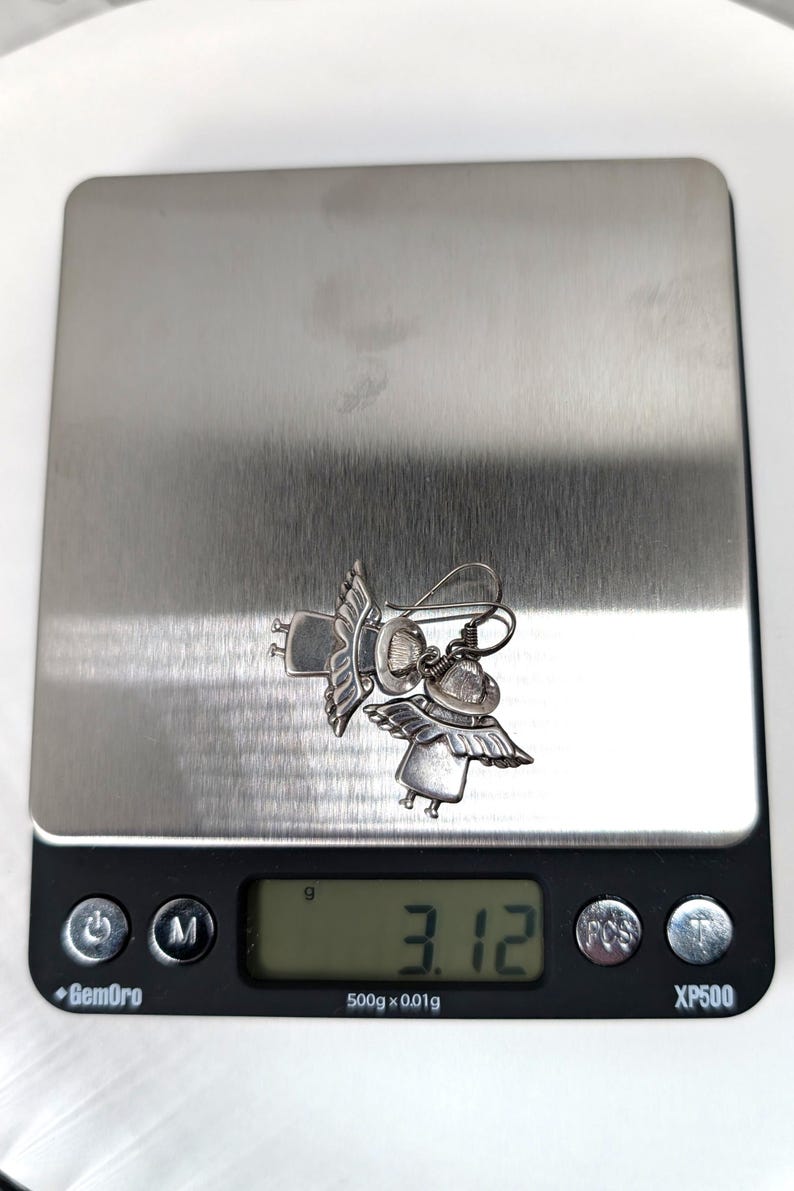 Op de afbeelding: Een paar zilverkleurige engelen oorbellen met gedetailleerde vleugels en hoeden, liggend op een digitale weegschaal. De weegschaal geeft een gewicht van 3,12 gram weer. De oorbellen zijn het middelpunt en tonen een grillig ontwerp.