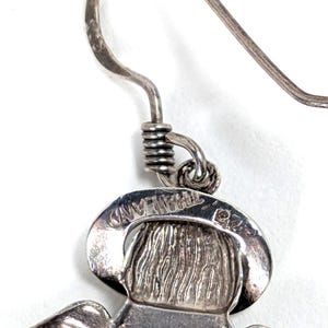 Op de afbeelding: Close-up van een zilverkleurige oorbel met een gedetailleerd ontwerp. De oorbel heeft een haak, een opgerolde draad en een hanger met een figuur met een hoed. Het woord "THAILAND" is zichtbaar op de hanger.