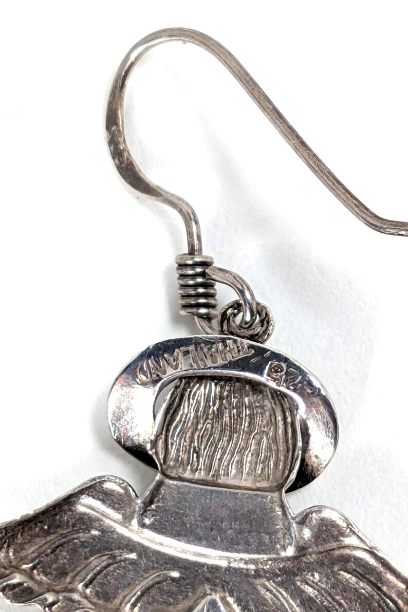 Op de afbeelding: Close-up van een zilverkleurige oorbel met een gedetailleerd ontwerp. De oorbel heeft een haak, een opgerolde draad en een hanger met een figuur met een hoed. Het woord "THAILAND" is zichtbaar op de hanger.