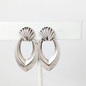 May include: A pair of silver earrings. Each earring has a seashell-shaped top and a teardrop-shaped, open-centre design. The earrings are displayed on a white stand against a white background.