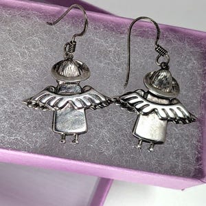 Op de afbeelding: Een paar zilverkleurige engelen oorbellen. Elke oorbel heeft een gestileerde engelenfiguur met een hoed, vleugels en een jurk. De oorbellen worden gepresenteerd in een paarse sieradendoos met een wit, getextureerd interieur.