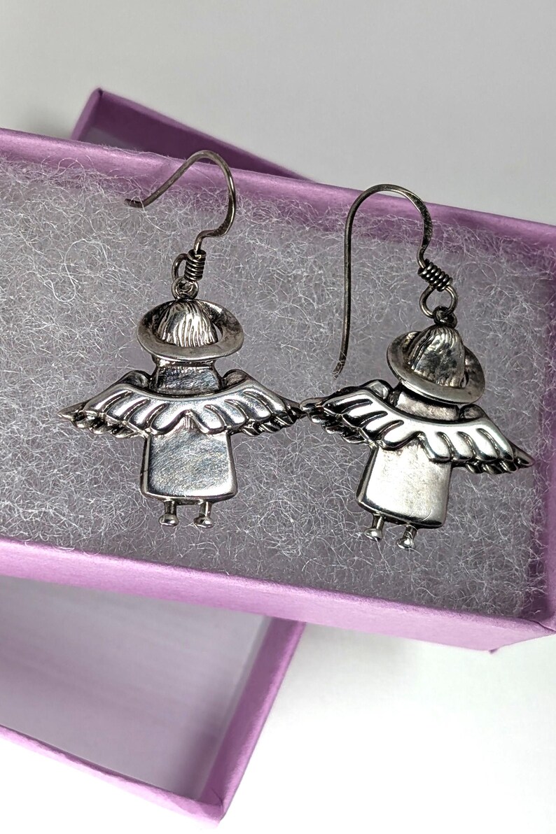 Op de afbeelding: Een paar zilverkleurige engelen oorbellen. Elke oorbel heeft een gestileerde engelenfiguur met een hoed, vleugels en een jurk. De oorbellen worden gepresenteerd in een paarse sieradendoos met een wit, getextureerd interieur.