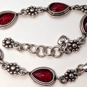 May include: Silver-toned bracelet featuring teardrop-shaped, deep red gemstones. The bracelet has a floral design between the gemstones and a heart-shaped clasp. A beautiful piece of jewelry.