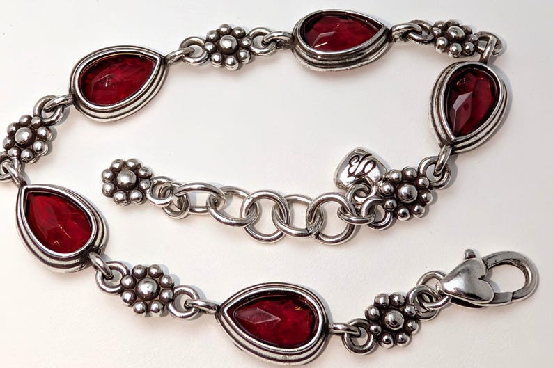 May include: Silver-toned bracelet featuring teardrop-shaped, deep red gemstones. The bracelet has a floral design between the gemstones and a heart-shaped clasp. A beautiful piece of jewelry.