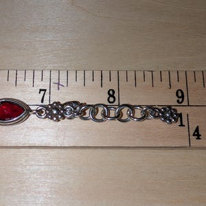May include: Silver-toned bracelet with a red, oval-shaped stone and floral accents. The bracelet is approximately 3 inches long, as measured by a ruler. The design includes a series of interconnected rings and decorative floral elements at each end.