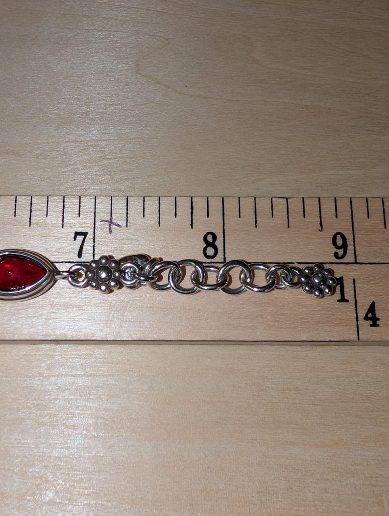 May include: Silver-toned bracelet with a red, oval-shaped stone and floral accents. The bracelet is approximately 3 inches long, as measured by a ruler. The design includes a series of interconnected rings and decorative floral elements at each end.
