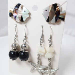 May include: A set of earrings displayed on a white card. The set includes two pairs of dangle earrings and two pairs of fish-shaped stud earrings. One pair has black and silver beads, the other has white shell pieces and silver starfish charms.