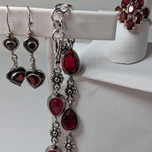 May include: A jewelry set featuring sterling silver earrings, a bracelet, and a ring, all adorned with vibrant red garnet gemstones. The earrings have heart-shaped designs, the bracelet has teardrop-shaped stones, and the ring has a cluster of garnets.