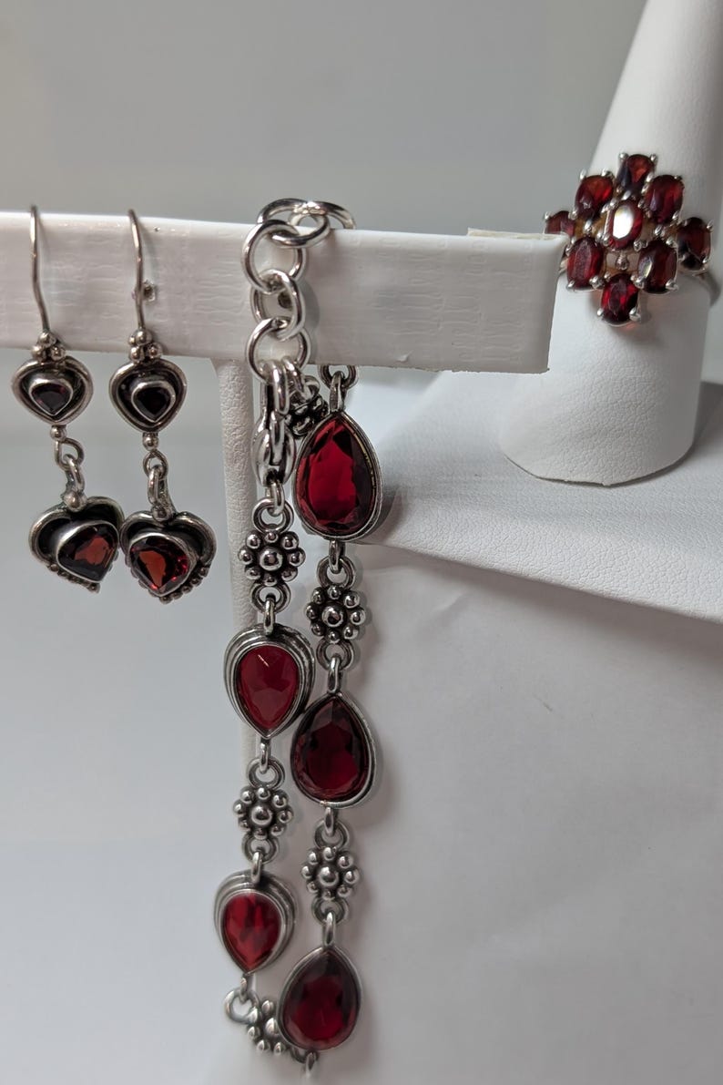May include: A jewelry set featuring sterling silver earrings, a bracelet, and a ring, all adorned with vibrant red garnet gemstones. The earrings have heart-shaped designs, the bracelet has teardrop-shaped stones, and the ring has a cluster of garnets.