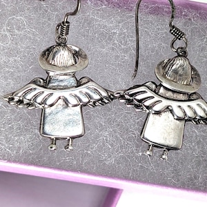 Op de afbeelding: Een paar zilverkleurige engelenoorbellen. Elke oorbel toont een gestileerde engel met een hoed, vleugels en kleine voetjes. De oorbellen worden gepresenteerd in een paarse sieradendoos met een gestructureerd, grijs interieur.