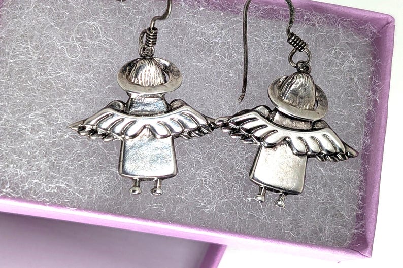 Op de afbeelding: Een paar zilverkleurige engelenoorbellen. Elke oorbel toont een gestileerde engel met een hoed, vleugels en kleine voetjes. De oorbellen worden gepresenteerd in een paarse sieradendoos met een gestructureerd, grijs interieur.