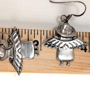 Op de afbeelding: Een paar zilverkleurige engel oorbellen. Elke oorbel heeft een engelontwerp met een hoed, vleugels en een jurk. De oorbellen worden getoond op een houten liniaal.