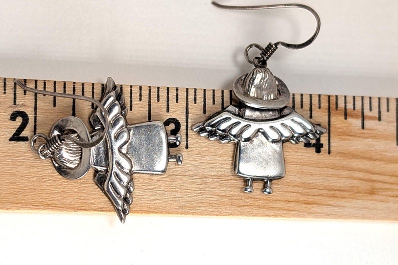 Op de afbeelding: Een paar zilverkleurige engel oorbellen. Elke oorbel heeft een engelontwerp met een hoed, vleugels en een jurk. De oorbellen worden getoond op een houten liniaal.