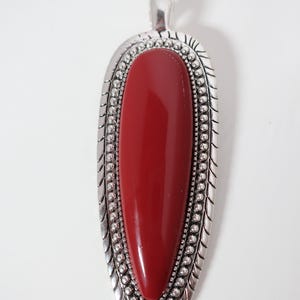 May include: A large, elongated pendant with a vibrant red center stone. The stone is encased in a silver-toned metal setting with a beaded border and decorative edges. The pendant has a loop at the top for a chain.