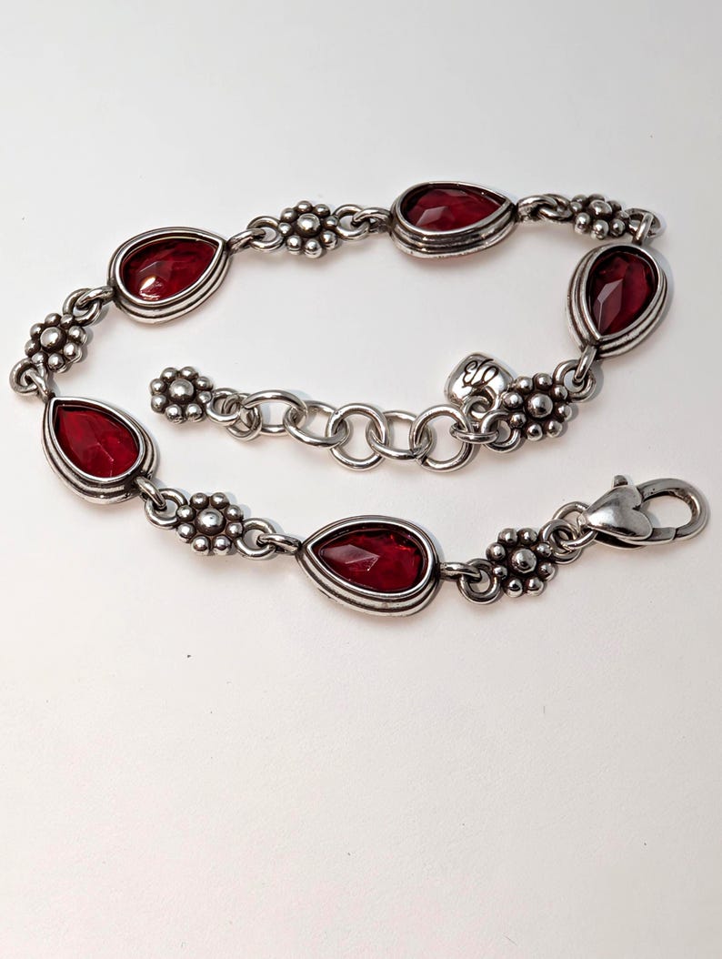 May include: Silver-toned bracelet featuring teardrop-shaped, deep red gemstones. The bracelet has a floral design between each gemstone and a secure clasp. A beautiful piece of jewelry.