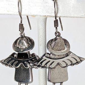 Op de afbeelding: Een paar zilverkleurige engelen oorbellen. Elke oorbel heeft een klein figuur met een hoed, vleugels en benen. De oorbellen hangen aan een gebogen draadhaak met een spiraalvormig detail.