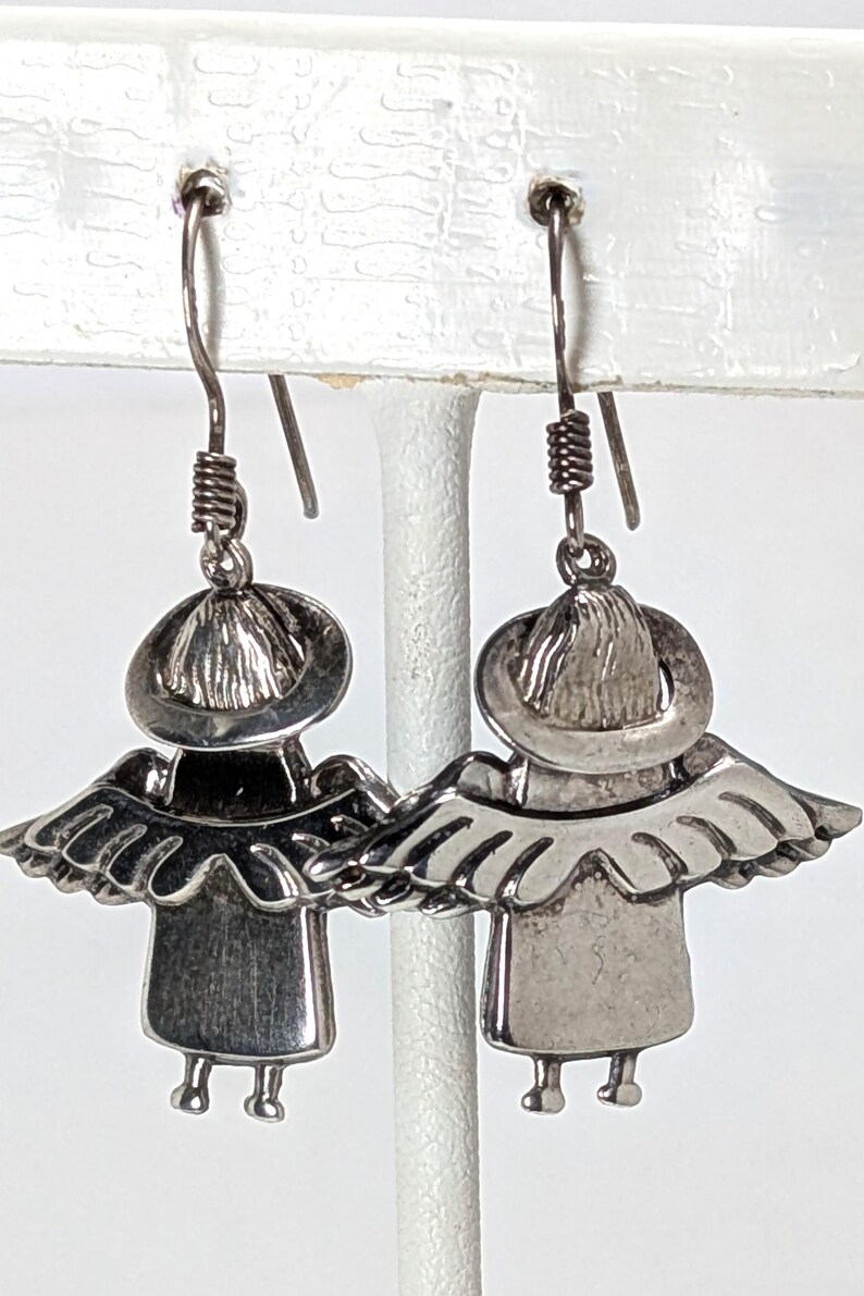 Op de afbeelding: Een paar zilverkleurige engelen oorbellen. Elke oorbel heeft een klein figuur met een hoed, vleugels en benen. De oorbellen hangen aan een gebogen draadhaak met een spiraalvormig detail.