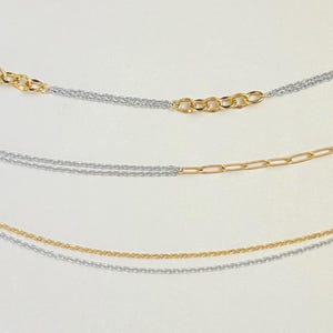 May include: Close-up of three delicate necklaces. Each necklace features a combination of silver and gold-toned chains. The top necklace has a chain of small gold rings. The middle necklace has a gold paperclip chain. The bottom necklace has a thin gold chain.