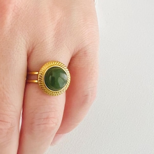 May include: A gold-colored ring with a large, round, dark green stone. The ring has a textured, rope-like border around the stone and a double band. The ring is on a finger against a white background.
