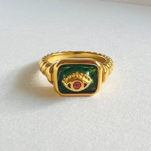 May include: Gold-toned ring featuring a square green stone with an eye design. The eye has a gold iris and a small pink center. The ring band has a textured, twisted rope design.
