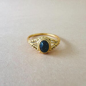May include: A gold-colored ring with an oval, dark blue stone speckled with glitter. The ring has an ornate design around the stone and on the band's shoulders. The ring is set against a light pink background.