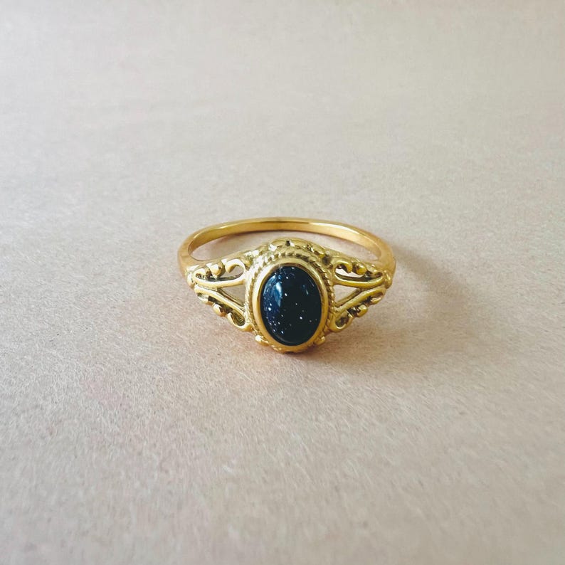 May include: A gold-colored ring with an oval, dark blue stone speckled with glitter. The ring has an ornate design around the stone and on the band's shoulders. The ring is set against a light pink background.