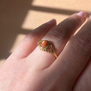 May include: A gold ring with an oval, orange gemstone. The ring has an ornate, filigree design around the gemstone and along the band. The ring is on a finger, with the background out of focus.