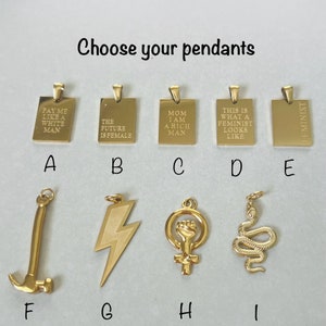 May include: Nine gold pendants with different designs and text. The pendants are arranged in two rows of five. The top row includes pendants with the text "Pay me like a white man", "The future is female", "Mom I am a rich man", "This is what a feminist looks like", and "Feminist". The bottom row includes pendants with a hammer, lightning bolt, female symbol with a fist, snake, and a blank pendant.