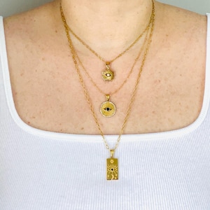 May include: A layered gold-tone necklace set featuring three distinct chains. The top chain has a small sunburst pendant, the middle chain features a circular evil eye charm with a central black stone, and the bottom chain holds a rectangular pendant.