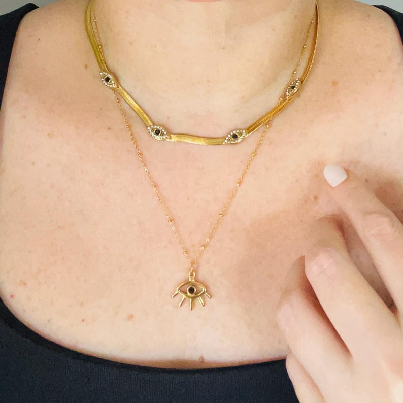 May include: A layered gold-tone necklace set featuring a flat herringbone chain adorned with three small eye-shaped charms with black centers and clear stones, and a delicate beaded chain with a single larger eye-shaped pendant with a black center.