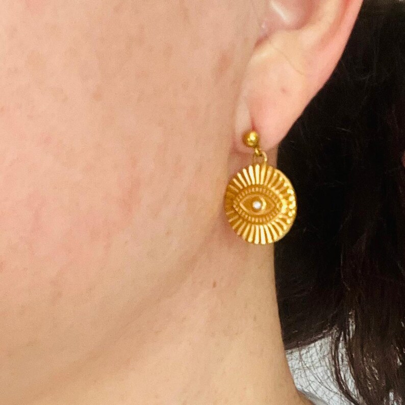Op de afbeelding: Gouden oorbellen met een rond, oogvormig ontwerp. Het oog heeft een wit centrum en is omgeven door een zonnestralenpatroon.