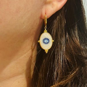 Op de afbeelding: Gouden oorbellen met een witte ovale hanger met een blauw boze oog ontwerp. De hanger is omgeven door een gouden rand en heeft een gouden ketting.